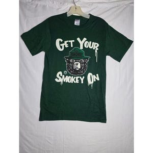 May include: A dark green short-sleeve t-shirt featuring a graphic of a bear wearing a green ranger hat. The text "GET YOUR SMOKEY ON" is printed in white above and below the bear illustration, with white paint-drip accents on the lettering.