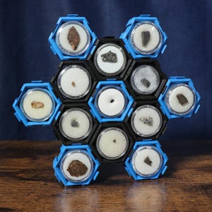 May include: A collection of small mineral or meteorite specimens housed in individual circular clear containers with white foam inserts, arranged within a modular blue and black hexagonal plastic display frame in a cluster formation.