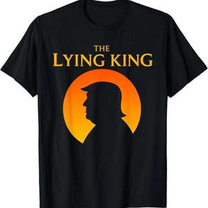 May include: A black short-sleeve t-shirt featuring a graphic of a person's silhouette in profile against a large orange sun. Above the silhouette, the text reads THE LYING KING in bold yellow capital letters. The design is centered on the chest.