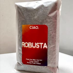May include: A rectangular bag of coffee beans in a semi-transparent white pouch. The label features a red-to-purple gradient with the text CIAO. and ROBUSTA in white. Additional text includes Origin: Kawi, Blitar, East Java, Variety: Tugusari, and Roastign Profile: Medium.