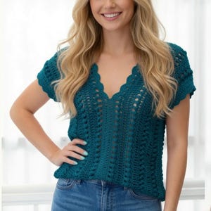 May include: A teal short-sleeved crochet top featuring a deep V-neckline with a scalloped edge. The garment is crafted with a vertical openwork stitch pattern and short sleeves with scalloped hems, paired with blue denim jeans.