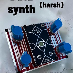 Op de afbeelding: Een rechthoekige Data Synth synthesizer met een zwart middenpaneel met een geometrische oogafbeelding, vier grote blauwe bedieningsknoppen en een rood en zilver gestreept metalen chassis. Het apparaat bevat labels voor USB 5V, output, pitch, disorder, confusion en Error Instruments.