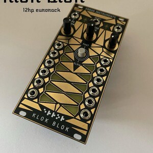 Puede incluir: Un módulo Eurorack negro y dorado con el texto "KLOK BLOK" y "ERROR INSTRUMENTS" impreso en él. El módulo tiene una perilla, un botón y una serie de conectores.