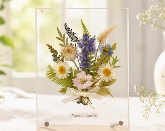 May include: A clear rectangular acrylic frame containing a pressed flower bouquet with white, blue, and green foliage, tied with a small white ribbon. The text Mima's Garden is printed at the bottom. The frame rests on a rectangular stone base.