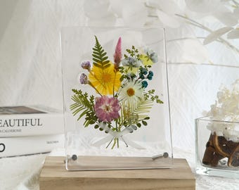 May include: A rectangular clear acrylic frame containing a pressed flower arrangement with yellow, pink, and white blossoms, green ferns, and blue accents. The flowers are tied with a small white ribbon and mounted on a rectangular wooden base.