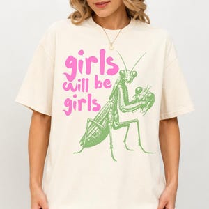 Op de afbeelding: Een off-white t-shirt met korte mouwen, voorzien van een afbeelding van een groene bidsprinkhaan en de roze tekst "girls will be girls" op de voorzijde. Het shirt heeft een ronde hals en een ruimvallend model met korte mouwen.