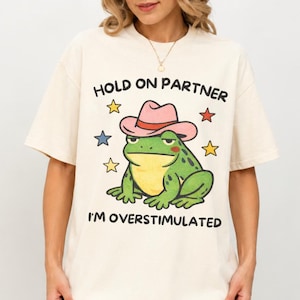 Peut inclure: Un t-shirt à manches courtes blanc cassé avec un motif de grenouille verte portant un chapeau de cowboy rose. Le texte "HOLD ON PARTNER" est inscrit au-dessus de la grenouille et "I'M OVERSTIMULATED" en dessous, entouré de petites étoiles.