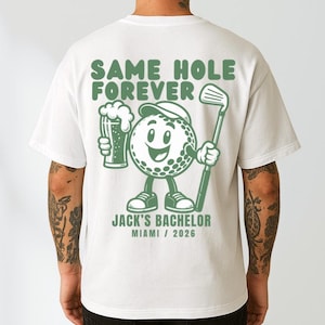 May include: A white t-shirt featuring a green graphic on the back. The design shows a cartoon golf ball wearing a cap, holding a beer mug and a golf club. Text reads: SAME HOLE FOREVER, JACK'S BACHELOR, MIAMI / 2026. The shirt has short sleeves.