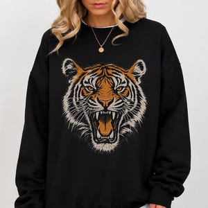 May include: A black long-sleeve sweatshirt featuring a large, detailed graphic of a roaring tiger head in shades of orange, black, and white. The tiger is centered on the chest, showcasing sharp teeth and intense eyes. The garment has a crew neckline.