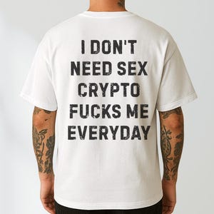 Op de afbeelding: Een wit T-shirt met korte mouwen en zwarte tekst in een verweerde stijl op de achterkant met de tekst: I DON'T NEED SEX CRYPTO FUCKS ME EVERYDAY. Het shirt heeft een ronde hals en een standaard pasvorm, getoond vanaf de achterkant.