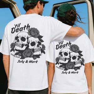 May include: Two white t-shirts featuring a black graphic print on the back. The design displays two skulls surrounded by roses, with the text 'til Death, the date 05.09.2026, and the names July & Mark printed in a bold, stylized black font.