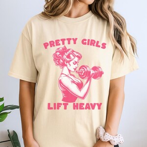 Könnte beinhalten: Ein cremefarbenes Kurzarm-T-Shirt mit einer rosa Grafik einer Person, die eine Hantel hebt. Der Text "PRETTY GIRLS" ist in Rosa über der Grafik gedruckt, und der Text "LIFT HEAVY" ist in Rosa unter der Grafik gedruckt.