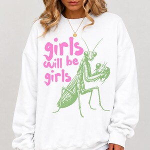 May include: A white long-sleeve sweatshirt featuring a green graphic illustration of a praying mantis and the pink text "girls will be girls" printed on the front. The garment has a crew neckline and long sleeves with ribbed cuffs and hem.