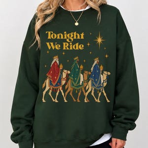 Könnte beinhalten: Ein dunkelgrünes Sweatshirt mit Rundhalsausschnitt und dem goldenen Schriftzug Tonight We Ride über einer Illustration von drei gekrönten Figuren in bunten Gewändern auf Kamelen, akzentuiert durch goldene Sterne auf dem dunkelgrünen Stoff.