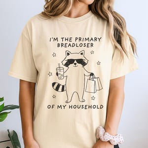 May include: A cream-colored short-sleeve t-shirt featuring a black line-art graphic of a raccoon wearing sunglasses, holding a drink and shopping bags. The text reads I'M THE PRIMARY BREADLOSER OF MY HOUSEHOLD, surrounded by small star illustrations.