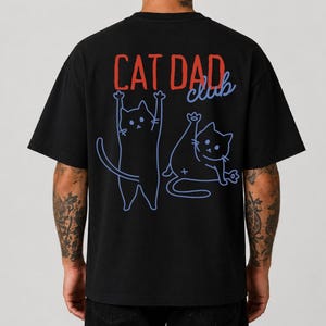 Könnte beinhalten: Ein schwarzes Kurzarm-T-Shirt mit einer Grafik auf der Rückseite. Das Design umfasst den roten Schriftzug CAT DAD und den blauen Schriftzug club, begleitet von zwei blauen Strichzeichnungen von Katzen in verspielten Posen auf dem Stoff.