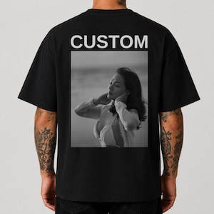 Può includere: Una t-shirt nera a maniche corte con la parola CUSTOM stampata in lettere maiuscole bianche sopra una stampa fotografica quadrata in bianco e nero di una persona che indossa un top chiaro con le mani vicino al viso.