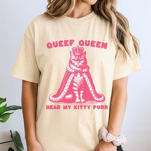 May include: A cream-colored short-sleeve t-shirt featuring a pink graphic of a cat wearing a crown and a royal cape. The text above the cat reads "QUEEF QUEEN" and the text below the cat reads "HEAR MY KITTY PURR" in a bold, pink, sans-serif font.