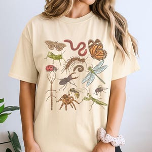 Peut inclure: Un t-shirt à manches courtes de couleur crème avec une impression graphique détaillée de divers insectes et arachnides, dont un papillon, une libellule, une coccinelle, une sauterelle, un scarabée, une araignée, un mille-pattes et un ver.
