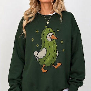 Peut inclure: Un sweat-shirt à manches longues vert forêt foncé arborant un motif central représentant un canard de dessin animé vêtu d'un costume de cornichon vert et bosselé. Le canard a des pattes orange et une aile blanche, entouré d'étoiles jaunes.