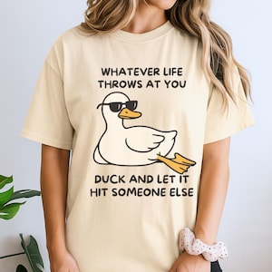 Puede incluir: Una camiseta de manga corta color crema con un gráfico de un pato blanco con gafas de sol negras. El texto sobre el pato dice WHATEVER LIFE THROWS AT YOU y el texto debajo dice DUCK AND LET IT HIT SOMEONE ELSE en fuente de color negro.