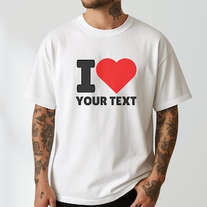 May include: A white short-sleeved t-shirt featuring a graphic print on the chest. The design consists of a black letter I, a large red heart symbol, and the words YOUR TEXT in bold black capital letters centred below the heart icon.