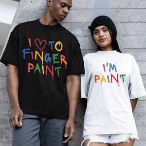 Könnte beinhalten: Ein schwarzes T-Shirt mit dem Text I LOVE TO FINGER PAINT in bunten, tropfenden Buchstaben und ein weißes T-Shirt mit dem Text I'M PAINT in bunten, tropfenden Buchstaben. Der Text ist in den Farben Rot, Blau, Gelb und Grün gehalten.