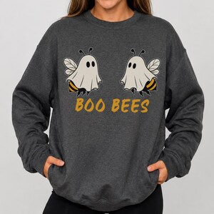 Può includere: Una felpa girocollo grigio antracite scuro con una grafica raffigurante due api che indossano costumi da fantasma bianchi. Sotto le api, la scritta BOO BEES è stampata in un audace colore giallo senape. La felpa ha maniche lunghe.