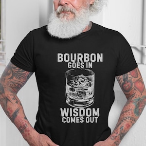 Può includere: Una t-shirt nera a maniche corte con una grafica bianca di un bicchiere pieno di cubetti di ghiaccio. Il testo sopra il bicchiere recita BOURBON GOES IN e il testo sotto il bicchiere recita WISDOM COMES OUT in un carattere stile blocco.