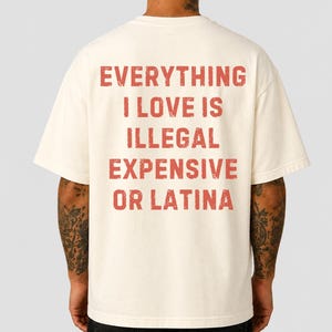 May include: An off-white short-sleeve t-shirt featuring red distressed-style text on the back that reads: EVERYTHING I LOVE IS ILLEGAL EXPENSIVE OR LATINA. The garment has a crew neckline and a relaxed fit, displaying the back view of the shirt.