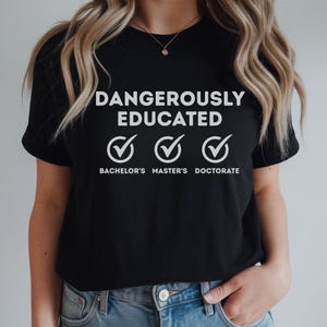 以下が含まれることがあります： 黒の半袖Tシャツ。胸元中央に白い文字で「DANGEROUSLY EDUCATED」とプリントされ、その下に3つのチェックマークアイコンが並んでいます。アイコンの下には「BACHELOR'S」「MASTER'S」「DOCTORATE」という白い文字が記載されています。