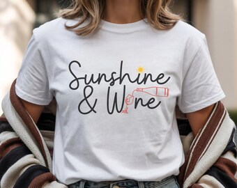 Puede incluir: Una camiseta blanca de manga corta con el texto negro Sunshine & Wine, un pequeño icono de sol amarillo sobre el texto y un dibujo lineal rojo de una botella de vino vertiendo en una copa integrado en la palabra Wine en el frente.