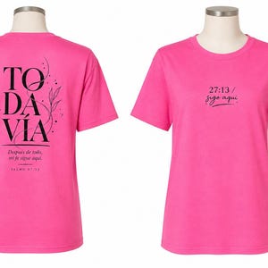 May include: A bright pink short-sleeved t-shirt featuring black text and botanical line art. The back displays the word TODAVÍA with the phrase Después de todo, mi fe sigue aquí and SALMO 27:13. The front shows 27:13 / sigo aquí.
