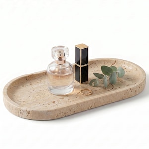 May include: An oval beige travertine stone tray with a porous, textured surface. Resting on the tray are a clear glass perfume bottle with a silver cap, a rectangular black lipstick tube with gold trim, a thin gold ring, and a small eucalyptus sprig.