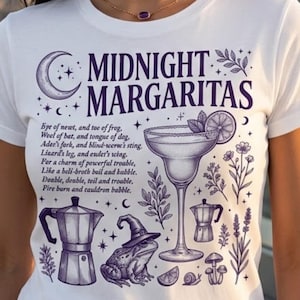 May include: A white short-sleeved t-shirt featuring a dark purple graphic print. The design includes the text Midnight Margaritas, a margarita glass, a frog wearing a witch hat, a coffee pot, and a poem excerpt, surrounded by botanical illustrations.