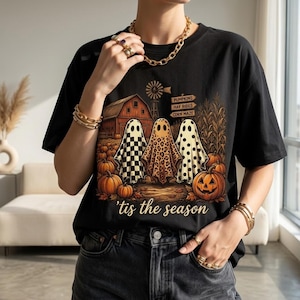 Puede incluir: Una camiseta negra con un gráfico de tres fantasmas frente a un granero, un molino y calabazas. Los fantasmas tienen patrones de cuadros, leopardo y lunares. Un letrero dice Pumpkins, Hay Rides, Corn Maze. El texto 'tis the season está debajo.