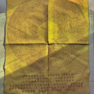 Op de afbeelding: Een rechthoekige goudkleurige stoffen banner met een groot cirkelvormig mandala-ontwerp bestaande uit dichte zwarte Chinese karakters. Onder de cirkel staat extra Chinese tekst in het zwart gedrukt. De stof heeft een subtiele glans en een zichtbare vouw.
