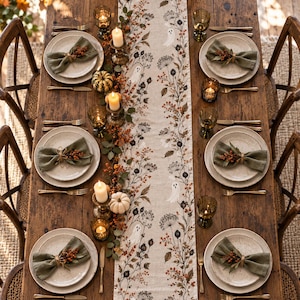 May include: A rustic wooden dining table set for four with a long beige table runner featuring a botanical print of ghosts, berries, and dried flowers. Each place setting includes a ceramic plate, a sage green napkin, and a small berry sprig.