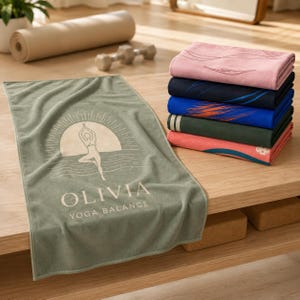 May include: A sage green rectangular yoga towel featuring a white graphic of a person in a tree pose against a sun and horizon line, with the text OLIVIA YOGA BALANCE printed below. A stack of five folded towels in various colors sits nearby.