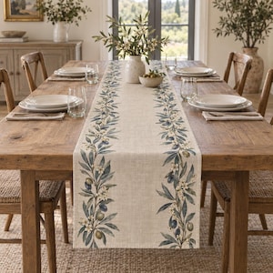 Puede incluir: Un camino de mesa rectangular de color beige con dos bordes verticales de ramas de olivo estampadas con hojas verdes, pequeñas flores blancas y aceitunas oscuras, colocado sobre una mesa de comedor de madera con platos blancos.