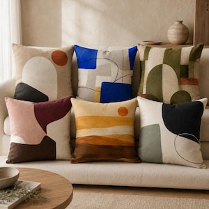 Könnte beinhalten: Eine Sammlung von sechs quadratischen Dekokissen, die auf einem Sofa angeordnet sind. Jedes Kissen zeigt ein eigenes abstraktes Design mit verschiedenen geometrischen Formen, Linien und Farbpaletten in Blau, Orange, Grün, Braun, Rosa und Creme.