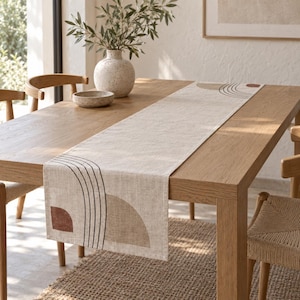 May include: A rectangular beige table runner featuring a printed design of black arched lines, a solid terracotta semi-circle, and a tan semi-circle. The runner is laid flat across a light brown wooden dining table with a textured surface.