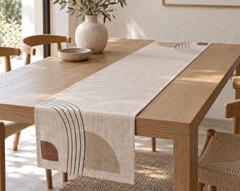 May include: A rectangular beige table runner featuring a printed design of black arched lines, a solid terracotta semi-circle, and a tan semi-circle. The runner is laid flat across a light brown wooden dining table with a textured surface.