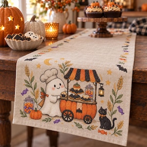 Peut inclure: Un chemin de table en lin beige orné d'une illustration imprimée représentant un fantôme en toque de chef près d'un chariot en forme de citrouille. Le motif inclut un chat noir, des épis de blé, des chauves-souris, des étoiles et des bonbons.
