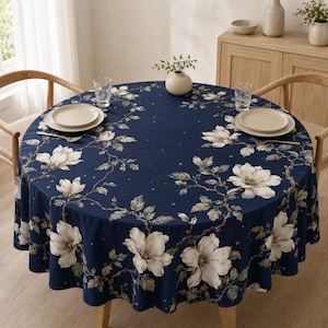Könnte beinhalten: Eine runde Tischdecke in tiefem Marineblau mit einem wiederkehrenden Muster aus großen weißen Magnolienblüten, grünen Blättern und kleinen verstreuten Sternen. Der Stoff liegt auf einem runden Tisch, der mit zwei weißen Tellern und zwei Gläsern gedeckt ist.