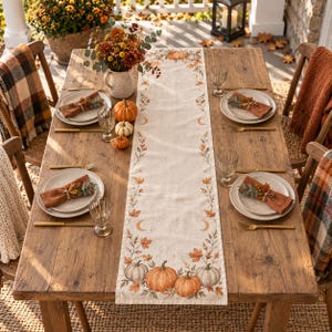 May include: A rectangular table runner featuring a printed pattern of orange pumpkins, autumn leaves, and crescent moons on a light beige fabric. The runner is centered on a wooden dining table set with plates, napkins, and glassware.
