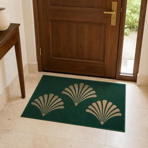 May include: A rectangular floor mat in a deep forest green color featuring three symmetrical, fan-shaped motifs in a light beige tone. The mat is positioned on a light-colored tile floor in front of a dark wood door with a gold-colored handle.