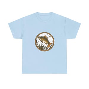 Może przedstawiać: Jasnoniebieski t-shirt z okrągłą grafiką na klatce piersiowej. Wzór przedstawia brązową rybę wyskakującą z wody, otoczoną obwódką w stylu liny, z widocznymi trzcinami i małymi bąbelkami wewnątrz okrągłej ramki.