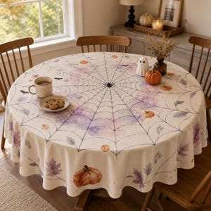 May include: A round tablecloth featuring a black spiderweb pattern over a watercolor background of purple and orange hues, decorated with small pumpkins, ghosts, and autumn leaves. A ceramic mug and a plate of cookies sit on the surface.