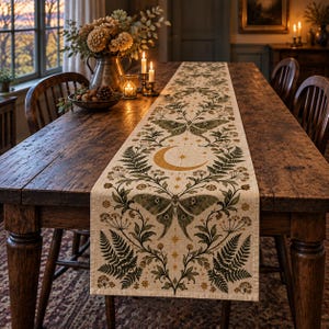 May include: A long, rectangular table runner made of light-colored fabric featuring a repeating botanical print. The design includes green moths, golden crescent moons, starbursts, and dark green fern fronds arranged in a symmetrical pattern.